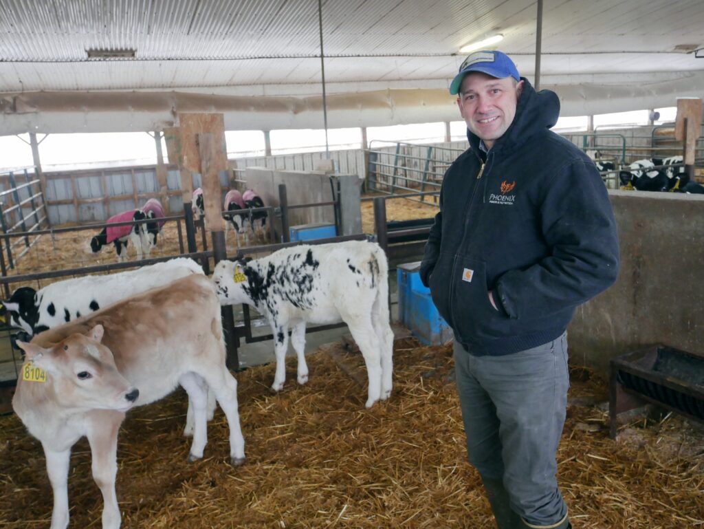 Kevin Kouri, a Nutritionist with Phoenix Feeds, stands in a calf barn, smiling at the camera. Behind him, young calves with ear tags rest in straw-bedded pens.